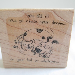 May include: A square wooden rubber stamp featuring a black ink illustration of a spotted dog chasing its tail. The text above the dog reads, "you did it! now, go chase your dream..." and the text below the dog reads, "or your tail or whatever..."