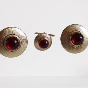 May include: A set of vintage gold-tone cufflinks and shirt studs. The set includes two circular cufflinks and a single shirt stud, each featuring a textured gold surface and a central, round, deep red stone. The jewelry is displayed against a white background.