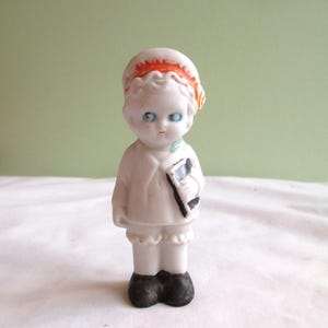 May include: A small, vintage porcelain doll with blue eyes, wearing a white outfit and a red-orange hat. The doll is holding a black and white book. The doll's shoes are black. The doll is standing on a white surface against a light green background.