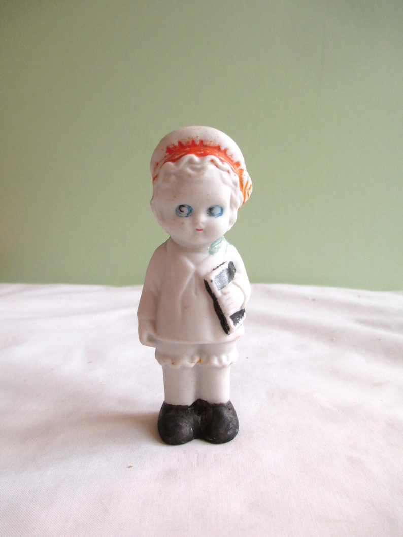 May include: A small, vintage porcelain doll with blue eyes, wearing a white outfit and a red-orange hat. The doll is holding a black and white book. The doll's shoes are black. The doll is standing on a white surface against a light green background.