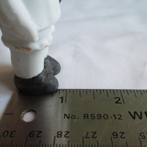 May include: A small, antique porcelain figurine, featuring a figure in white clothing and black shoes. The figurine is measured with a metal ruler, showing a scale in inches. The figure's legs and feet are visible, with a focus on the details of the clothing and footwear.