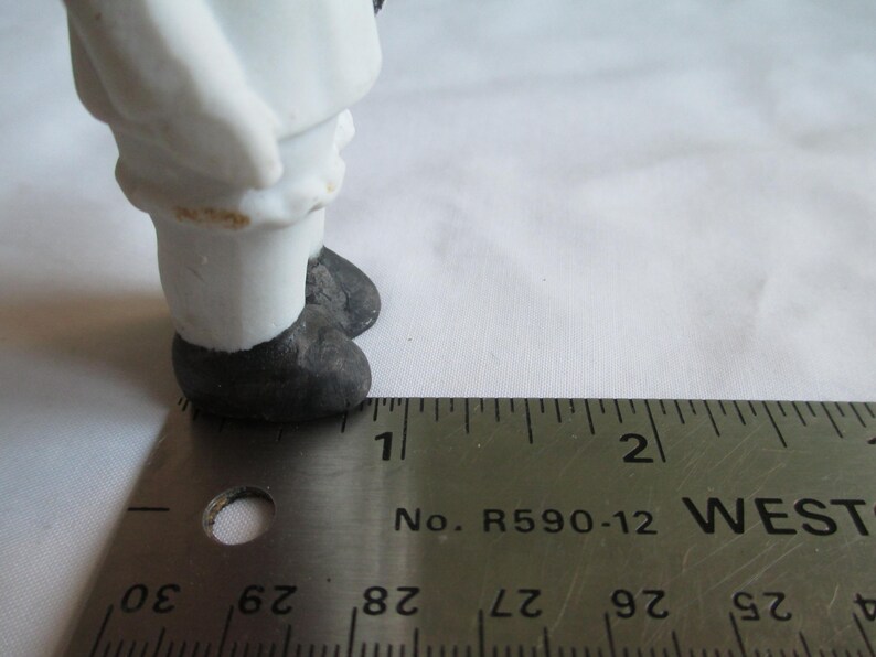 May include: A small, antique porcelain figurine, featuring a figure in white clothing and black shoes. The figurine is measured with a metal ruler, showing a scale in inches. The figure's legs and feet are visible, with a focus on the details of the clothing and footwear.