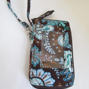 May include: A quilted fabric wristlet wallet in a dark brown, light blue, and teal floral paisley pattern. The wallet features a clear plastic ID window on the front, a dark brown zipper closure with a fabric pull, and a detachable matching wrist strap.