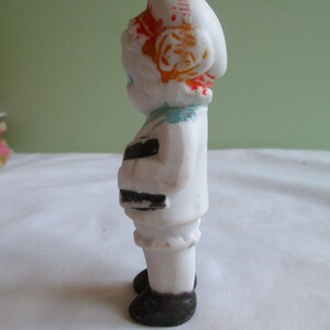 May include: A small, vintage porcelain figurine of a person in a white outfit with black accents. The figurine has a white hat with orange and red details, blue eyes, and black shoes. The figure is standing upright.