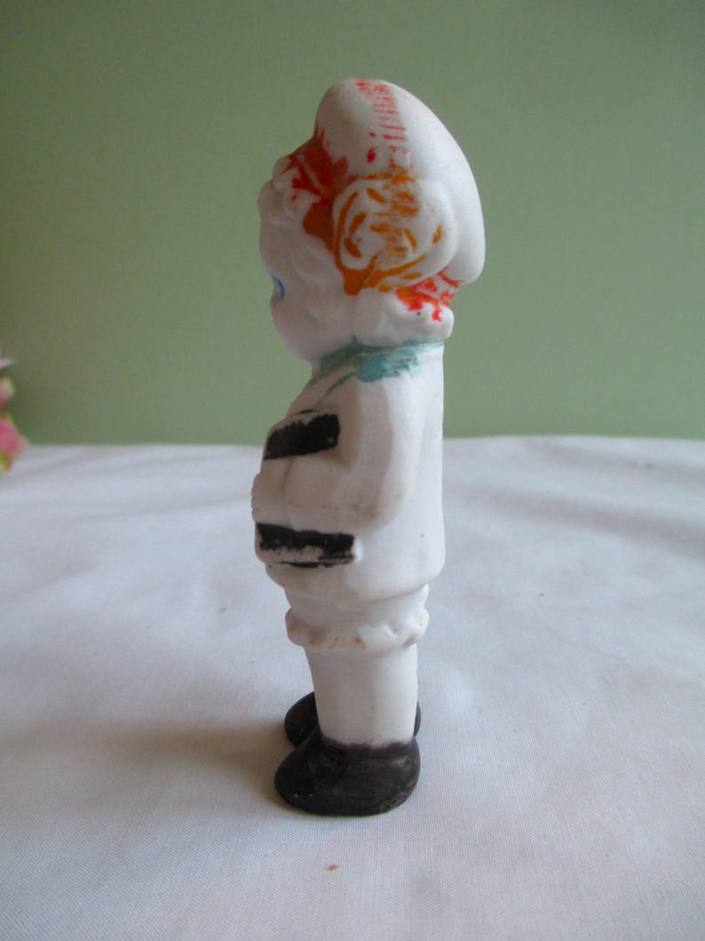 May include: A small, vintage porcelain figurine of a person in a white outfit with black accents. The figurine has a white hat with orange and red details, blue eyes, and black shoes. The figure is standing upright.