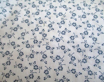 May include: Close-up of a white fabric with a repeating pattern of small, navy blue floral sprigs. The delicate flowers and leaves are evenly distributed across the surface, creating a classic, vintage aesthetic. The fabric appears to be cotton or a similar lightweight material.