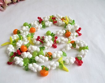 May include: A decorative garland featuring white cloud-like beads, interspersed with colorful glass fruit and leaves. The garland includes orange, green, and red fruit, along with yellow and green leaves. The garland is arranged on a white surface.