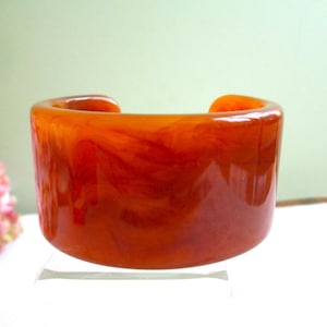 Op de afbeelding: Een brede, oranjerode manchetarmband met een gemarmerd effect. De armband heeft een rechthoekige vorm met afgeronde randen en een gladde, glanzende afwerking. De kleur is een warme, amberkleurige tint, met kleurvariaties.