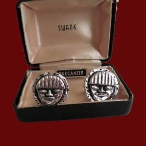 Può includere: Un paio di gemelli d'argento in una scatola nera con un'etichetta bianca che dice "Swank Buccaneer". I gemelli presentano un design di viso stilizzato.