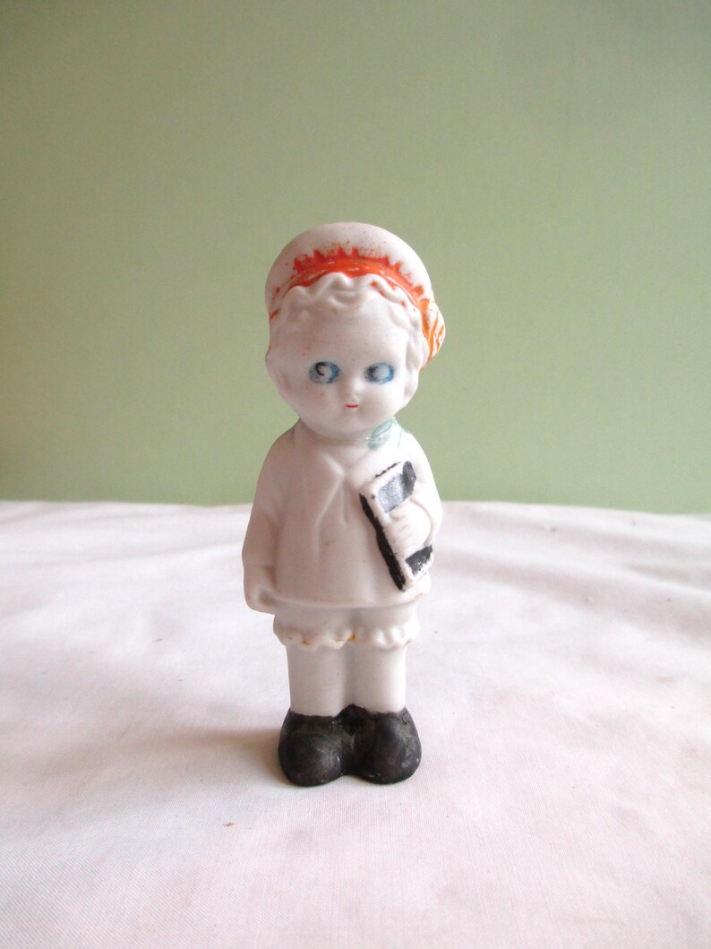 May include: A small, vintage porcelain doll with blue eyes, wearing a white outfit and a red-orange hat. The doll is holding a black and white book. The doll's shoes are black. The doll is standing on a white surface against a light green background.