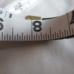 May include: A white measuring tape with black markings, showing the numbers 6, 8, and 2. The tape is partially rolled up and the end is secured with a metal clasp.