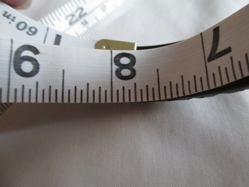 May include: A white measuring tape with black markings, showing the numbers 6, 8, and 2. The tape is partially rolled up and the end is secured with a metal clasp.