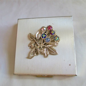 May include: A silver compact with a floral design featuring a cluster of colorful rhinestones. The compact has a hinged lid and a mirror inside.