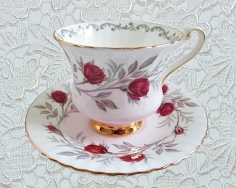 May include: A white teacup and saucer with a pink floral pattern featuring red roses and gray leaves. The teacup has a gold rim and a gold band around the base.