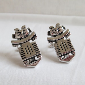 May include: A pair of silver cufflinks featuring a stylised design of a human figure. The cufflinks are detailed with a face and geometric patterns.