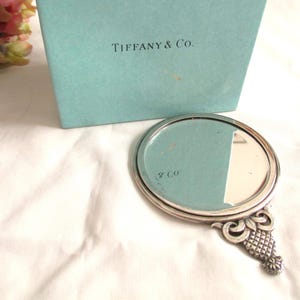 May include: A silver-toned hand mirror with a round mirror and ornate handle. The mirror is next to a Tiffany & Co. box. The handle has a decorative design. The mirror is on a white surface.