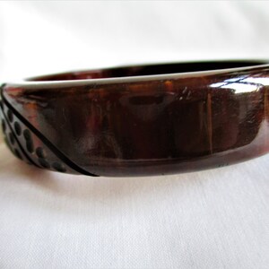 May include: A dark brown, glossy, plastic bangle bracelet with a geometric design on one side. The design features a series of raised circles.