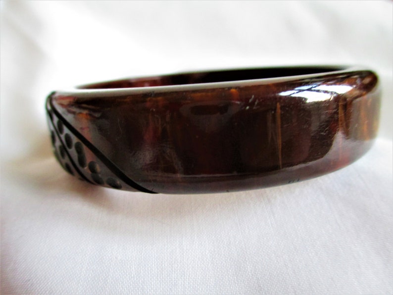 May include: A dark brown, glossy, plastic bangle bracelet with a geometric design on one side. The design features a series of raised circles.