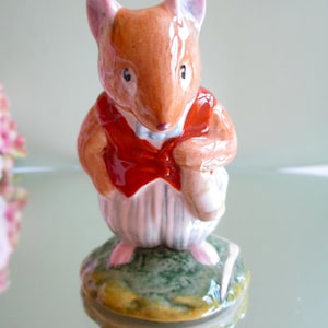 Peut inclure: Figurine en céramique représentant une souris vêtue d'un gilet rouge et d'une chemise blanche à rayures verticales. La souris est posée sur un socle vert et jaune. La souris a une fourrure brune et des oreilles roses.