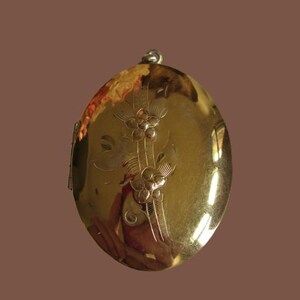 May include: A gold locket with a floral design engraved on the front. The locket has a hinged opening and a loop for attaching a chain.