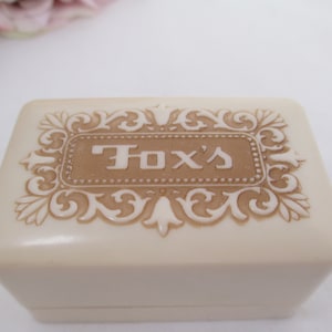 May include: A small, off-white, rectangular box with a raised, ornate design in brown. The box has the words "Fox's" embossed in brown on the top.