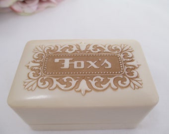 May include: A small, off-white, rectangular box with a raised, ornate design in brown. The box has the words "Fox's" embossed in brown on the top.
