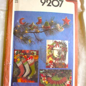 May include: Simplicity 9207 sewing pattern featuring Christmas-themed decorations. The pattern includes designs for stockings, a wreath, tree skirt, and ornaments. The cover displays colorful illustrations of the finished projects, with a blue background and a brown border.