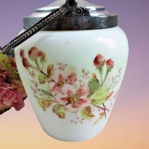 Puede incluir: Un tarro de cristal blanco con un diseño floral pintado a mano que presenta flores rosas y hojas verdes. El tarro tiene una tapa de plata metálica y un asa giratoria de plata a juego en la parte superior como recipiente decorativo.