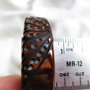 May include: A brown and black bakelite bangle bracelet with a geometric design. The bracelet is 1 inch wide and is being measured with a ruler.