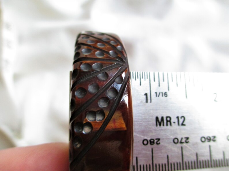 May include: A brown and black bakelite bangle bracelet with a geometric design. The bracelet is 1 inch wide and is being measured with a ruler.