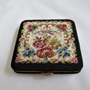 May include: A vintage, square compact with a black frame and a needlepoint floral design. The needlepoint features a variety of flowers in shades of red, pink, blue, and white, set against a cream background. The compact has a gold-colored clasp.