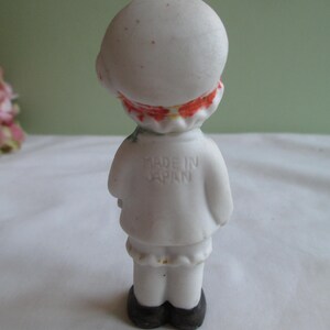 May include: A small, vintage figurine of a person, viewed from the back. The figurine is primarily white with red and orange accents around the neck and on the head. The figurine has black shoes and the text "MADE IN JAPAN" is visible on the back.