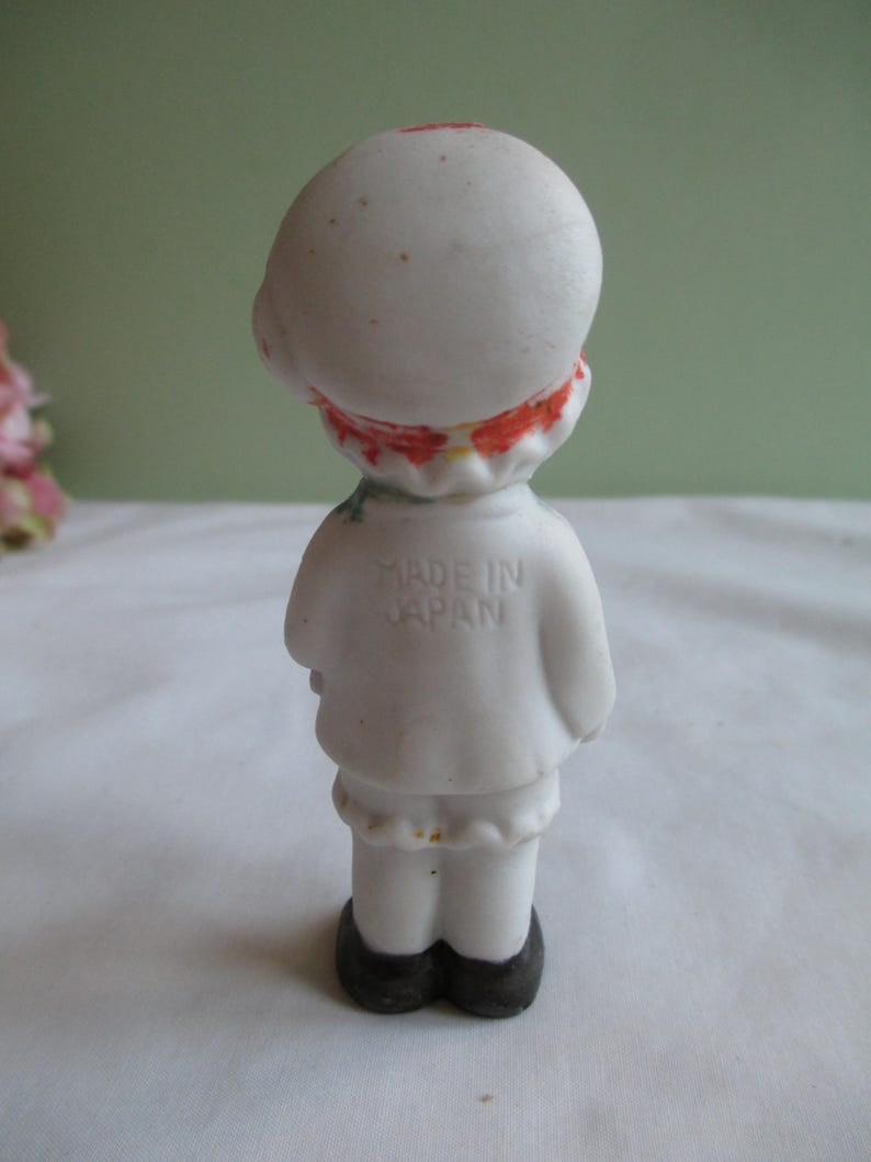 May include: A small, vintage figurine of a person, viewed from the back. The figurine is primarily white with red and orange accents around the neck and on the head. The figurine has black shoes and the text "MADE IN JAPAN" is visible on the back.