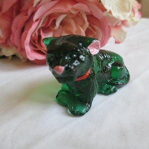 Puede incluir: Pequeña figura de gato de cristal verde esmeralda con collar rojo y detalles rosas en orejas y nariz. El gato está tumbado, con flores rosas y blancas de fondo.