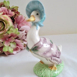 Puede incluir: Una figurita de porcelana de un pato con un gorro azul y un vestido morado y verde. El pato está de pie sobre una base verde que parece hierba.