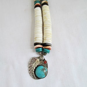 May include: A handcrafted necklace with a pendant featuring a turquoise stone and a small red stone, with a silver leaf design. The necklace has a double strand of white, disc-shaped beads, with turquoise, brown, and black accent beads.