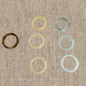May include: A collection of seven circular rings arranged in a grid. The rings feature a smooth, polished finish and come in various translucent colors, including dark brown, pale yellow, light green, soft blue, and a milky white shade.