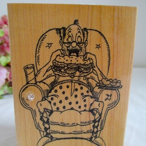 Op de afbeelding: Houten stempel met een cartoon illustratie van een man in een fauteuil, die meerdere hamburgers eet. De man draagt een bril en heeft een verbaasde uitdrukking. Een bord met eten en een drankje zijn ook afgebeeld. Een kleine edelsteen is aan de zijkant bevestigd.