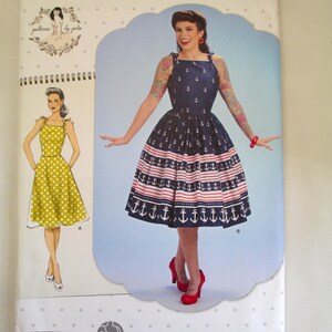 May include: Simplicity sewing pattern S8873 featuring a vintage-style dress with an anchor print. The dress has a navy blue bodice and a red and white striped skirt. The pattern includes a drawing of the dress and the Simplicity logo.