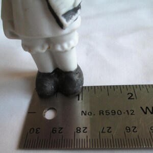 May include: A porcelain figurine's lower half is visible, featuring white legs, a ruffled hem, and black shoes. A metal ruler, marked in inches, is placed next to the figurine for scale. The figurine's design includes black accents.