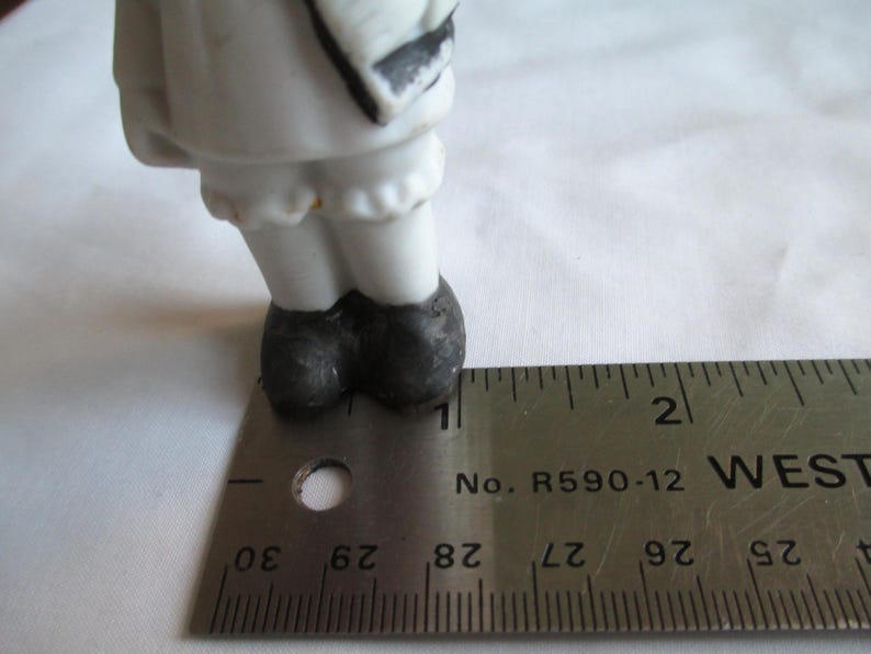 May include: A porcelain figurine's lower half is visible, featuring white legs, a ruffled hem, and black shoes. A metal ruler, marked in inches, is placed next to the figurine for scale. The figurine's design includes black accents.