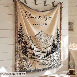May include: A beige woven throw blanket with a mountain and forest landscape design. The blanket has a fringed edge and features the names "Olivia & Liam" and the date "June 15, 2026" in black script.