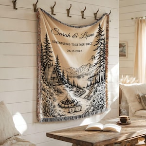 May include: Beige woven throw blanket with a black and white illustrated landscape scene. The scene includes a cabin, trees, mountains, and a lake. The blanket has the text "Sarah & Liam" and "Adventuring Together Since 06.15.2026".