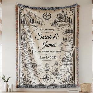 May include: A woven tapestry featuring a map design with mountains, a castle, and a lake. The map includes the text "The Journey of Sarah & James" and the date "June 15, 2026". The tapestry has a cream background with black and brown details and fringe.