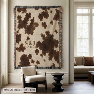Personalized cow print woven blanket with custom initials and established year, brown and cream western pattern, rustic farmhouse wall decor, fringe edges, and cozy country couple keepsake style.
