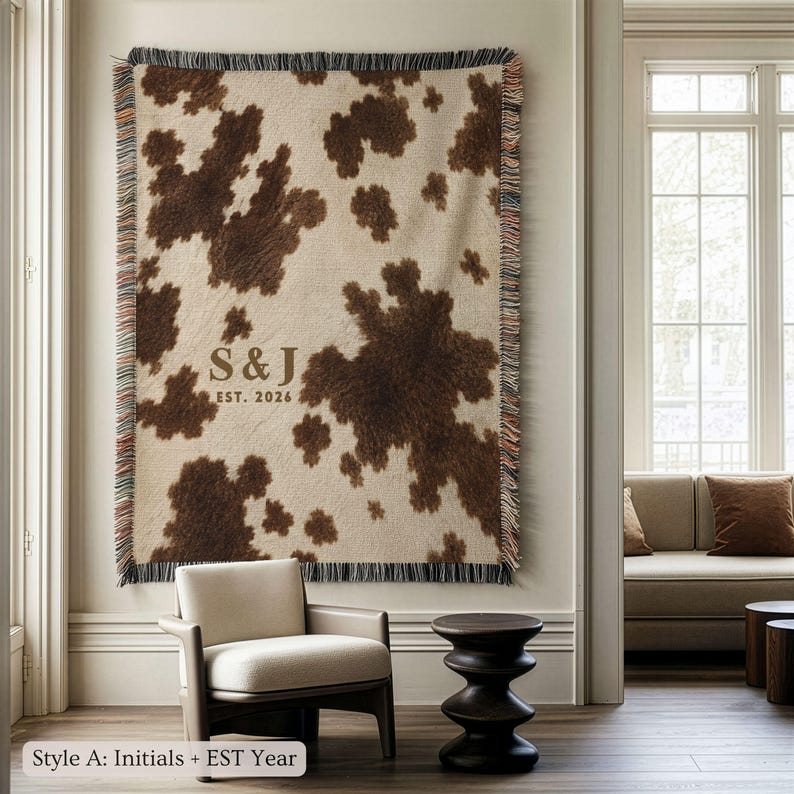 Personalized cow print woven blanket with custom initials and established year, brown and cream western pattern, rustic farmhouse wall decor, fringe edges, and cozy country couple keepsake style.
