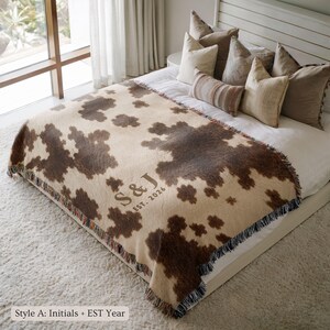 Personalized cow print woven blanket with custom initials and established year, brown and cream western pattern, rustic farmhouse wall decor, fringe edges, and cozy country couple keepsake style.