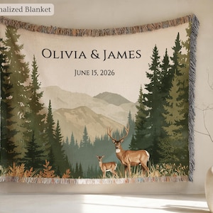 May include: A personalized blanket featuring a forest scene with deer and mountains. The blanket has the names "OLIVIA & JAMES" and the date "JUNE 15, 2026" printed on it. The blanket has a fringed edge and the words "Personalized Blanket" in the top left corner.
