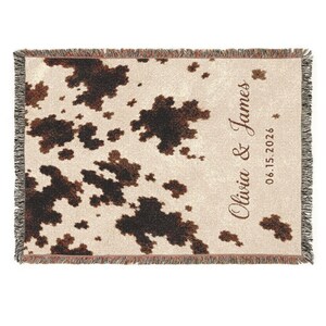 Personalized cow print woven blanket with custom couple names and wedding date, brown and cream western pattern, rustic farmhouse wall decor, fringe edges, and cozy country wedding keepsake style.