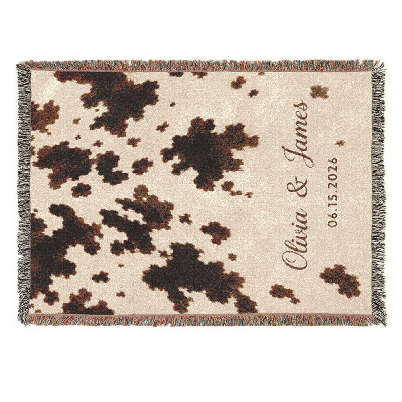 Personalized cow print woven blanket with custom couple names and wedding date, brown and cream western pattern, rustic farmhouse wall decor, fringe edges, and cozy country wedding keepsake style.